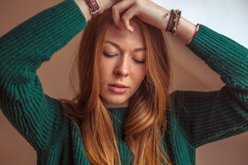 Portrait d’une femme rousse en pull vert, les bras relevés au-dessus de la tête.