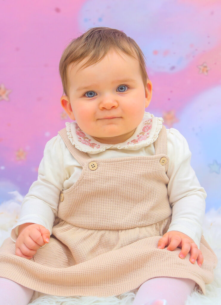 Bébé assis en robe beige devant un fond rose et bleu au studio.