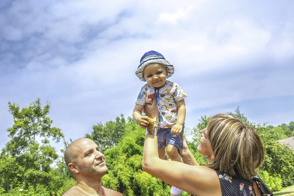 Un jeune enfant coiffé d’un chapeau, porté par ses parents dans un jardin.
