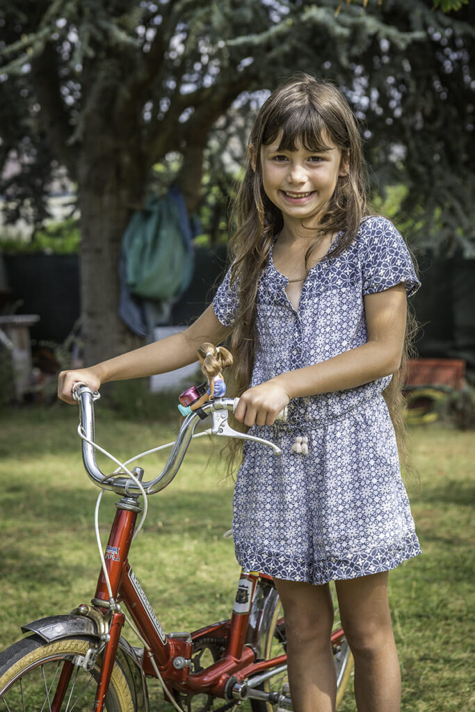 Enfant souriante tenant un vélo rouge dans un jardin.